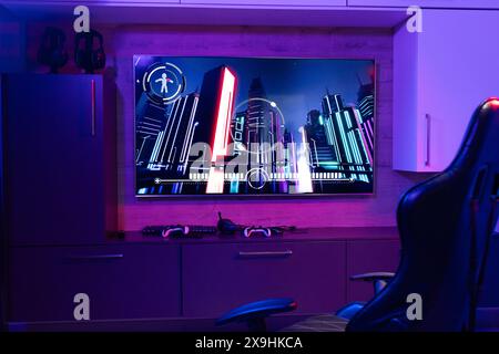 At home, facing neon cityscape art on a purple glow, seated in a gaming chair Stock Photo