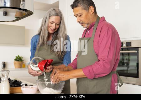 At home, diverse senior couple, both wearing aprons, baking together and smiling. Caucasian wife with long gray hair pouring flour, while biracial hus Stock Photo