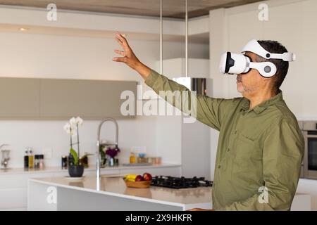 At home, biracial senior male in modern kitchen wearing virtual reality headset, copy space. Reaching out with one hand, interacting with virtual envi Stock Photo