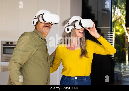 At home, diverse senior couple wearing virtual reality headsets in modern kitchen. Smiling and looking around, appearing to enjoy virtual experience t Stock Photo