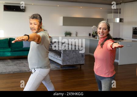 At home, diverse senior couple practicing yoga together in modern living room. Mature Caucasian woman with long gray hair and senior biracial man with Stock Photo