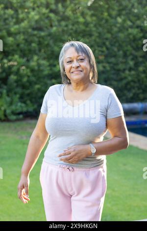 Outdoors, biracial senior woman smiling while standing in casual attire. She is in garden with lush greenery and a pool in background, enjoying serene Stock Photo