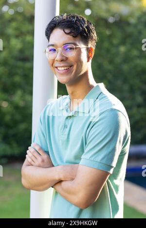 Young biracial man standing outdoors, smiling with arms crossed. Greenery background adding serene touch to scene, creating calm and peaceful atmosphe Stock Photo