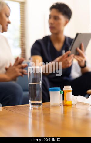 At home, young biracial male nurse showing tablet and medicine to senior female patient. They are in cozy living room with medication and a glass of w Stock Photo