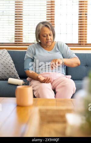 At home, biracial senior woman checking her pulse with blood pressure monitor. Sitting on blue sofa in bright living room with wooden blinds and cozy Stock Photo