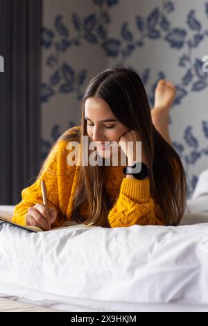 At home, young Caucasian woman relaxing on bed, writing in notebook with smile. Floral wallpaper and cozy bedding creating a serene home spa atmospher Stock Photo