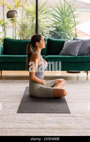 At home, young Caucasian woman meditating on yoga mat in casual workout clothes. Bright living room with green background, green sofa, and large windo Stock Photo