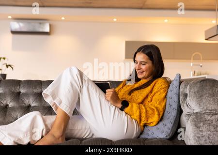 At home, young Caucasian woman relaxing on couch, smiling while using tablet. Modern living room with soft lighting and contemporary decor, creating c Stock Photo