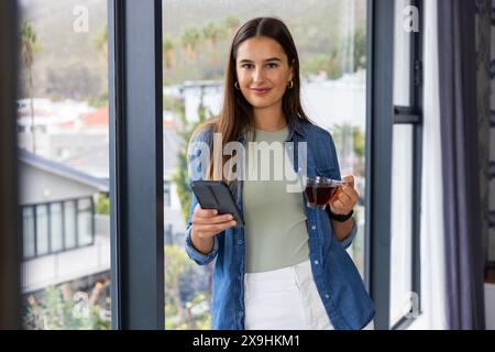 At home, young Caucasian woman holding smartphone and cup of tea, smiling at camera. She is standing by large window with view of suburban neighborhoo Stock Photo