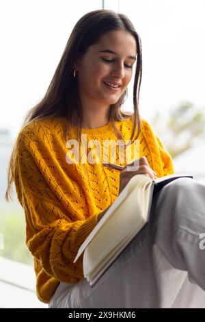 At home, young Caucasian woman in yellow sweater writing in a notebook Stock Photo