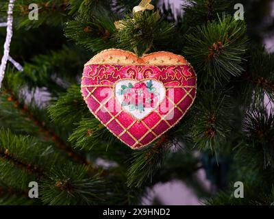 A cross stitched Christmas Timeless Elegance ornament on to a Christmas tree. This heart shape Christmas ornament with red flowers embroidered and mad Stock Photo