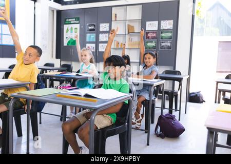 Diverse group of children raising hands eagerly in a school classroom. They are engaged in a learning activity, showcasing enthusiasm and curiosity. Stock Photo