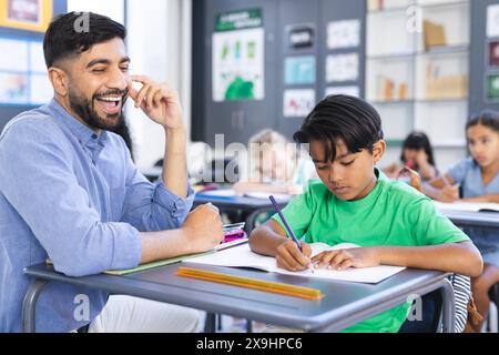 A young Asian male teacher smiles, guiding biracial boy in writing Stock Photo