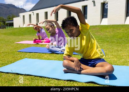 Biracial children practice yoga outdoors, with a boy in yellow leading the stretch Stock Photo