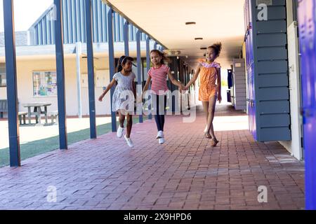 Three biracial girls are holding hands and walking through a school corridor with copy space. They appear joyful, with the school lockers and notice b Stock Photo