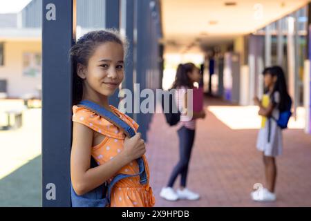 Biracial girl with a backpack smiles, standing in a school corridor with copy space Stock Photo