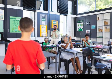 In school, young Asian male teacher and diverse young students are learning. They are focusing on a recycle poster and math signs, unaltered Stock Photo