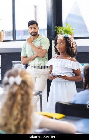In school, Asian male teacher watches as biracial female student presents Stock Photo