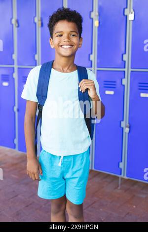 A biracial boy with a bright smile stands in front of blue lockers in school Stock Photo