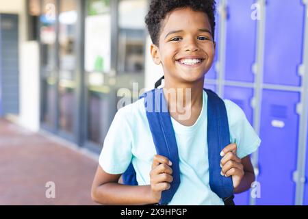 Biracial boy with a bright smile stands in front of lockers at school, wearing a blue backpack. His curly hair and casual school attire suggest a posi Stock Photo