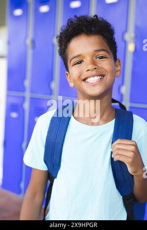 Biracial child with a bright smile stands in front of blue lockers at school. Wearing a light blue t-shirt and backpack, he exudes confidence and happ Stock Photo