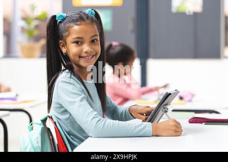 In school, young biracial girl holding tablet smiles at camera in classroom. Another young girl sits behind her, focusing on her own tablet, learning, Stock Photo