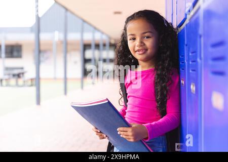 In school, outdoors, young biracial girl holding a book stands by lockers, copy space Stock Photo