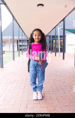 In school, outdoors, a biracial young girl holding a tablet stands smiling Stock Photo