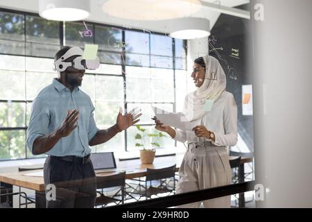 In modern business office, African American man in VR headset, part of diverse team. An indian female team leader holding documents, discussing ideas Stock Photo