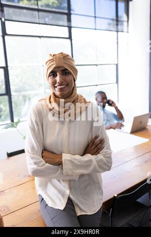 In modern business office, diverse team with young woman in beige headscarf standing. African American male colleague in background talking on smartph Stock Photo