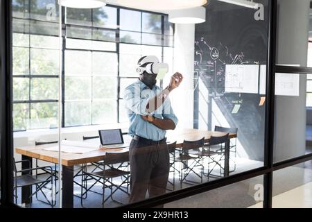 Young African American man in business office using VR headset, engaging with virtual screen. Standing near large window, with a laptop on a table in Stock Photo