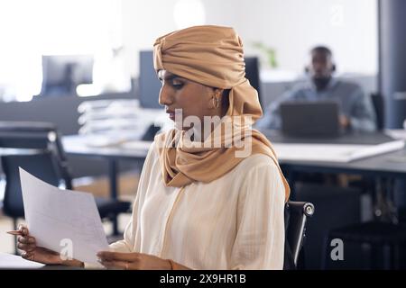 At modern business office, indian woman in beige headscarf reads document. In background, African American man working on laptop at a desk, unaltered Stock Photo