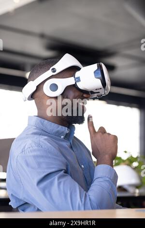 Young African American male wearing virtual reality headset in business office. He has short black hair, beard, and is smiling while touching headset Stock Photo