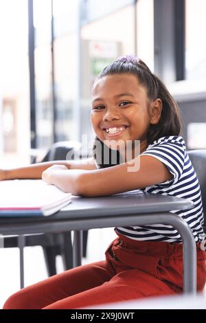 In school, young biracial girl with a bright smile sitting at a desk in a classroom Stock Photo