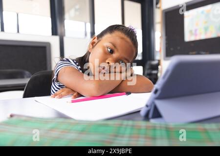 In school, young biracial girl with dark hair resting on desk, looking at tablet in classroom Stock Photo