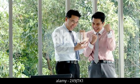 Success Businessman happy got good news from mobile smart phone that business so success. Asian young business man raise hand with happiness, smiling, Stock Photo