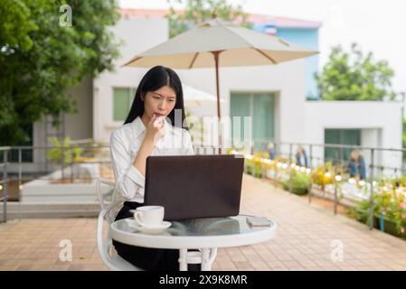 A focused woman working on her laptop at an outdoor terrace cafe with coffee and trees in the background Stock Photo