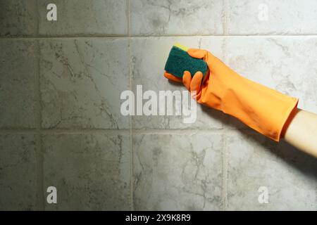 A hand in an orange latex glove brings a sponge to a tiled wall: disinfection and cleaning in the apartment. Stock Photo