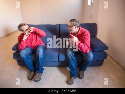 A middle-aged man in a red shirt and jeans sits on a couch, arguing with himself. Stock Photo