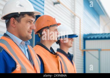 Three builders in helmets and vests stand in row. Friendly construction team of Caucasian appearance. Portraits of young workers at construction site on summer day. Stock Photo