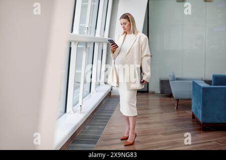 Portrait of beautiful pregnant businesswoman confidently working on laptop in a modern business office with a light blue background, wearing a stylish Stock Photo