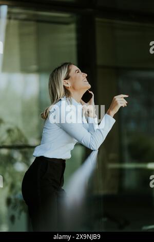 A smiling woman in a light blue shirt leans on a railing, holding her phone, outside a modern office building, relishing a moment of relaxation. Stock Photo
