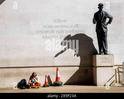 London, UK. 2nd June, 2024. A woman relaxes in the sunshine under a shadow cast by the statue of George Orwell. The statue of George Orwell by the British sculptor Martin Jennings was unveiled on 7 November 2017 outside Broadcasting House, the headquarters of the BBC, in London. The wall behind the statue is inscribed with George Orwell's words If liberty means anything at all, it means the right to tell people what they do not want to hear. This was a quote from an unused preface to Animal Farm. Credit: Mark Thomas/Alamy Live News Stock Photo