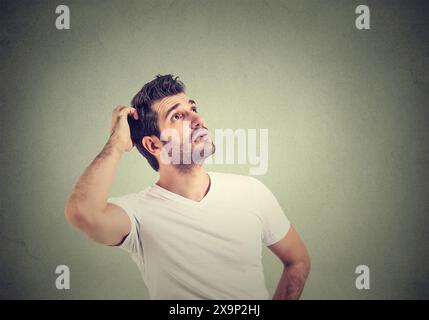 Young man in white t-shirt having doubts thinking while scratching head isolated on isolated gray wall background Stock Photo