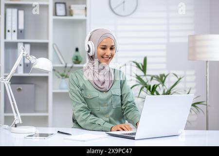Smiling woman in hijab wearing headphones, working on a laptop in a modern office. Professional workspace with plants and white decor. Stock Photo