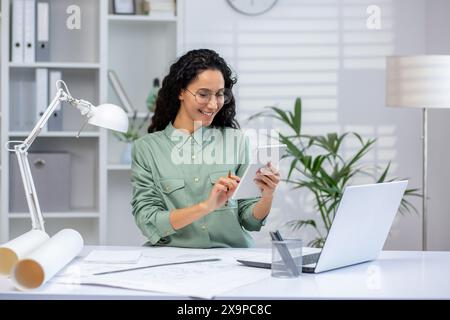 Smiling woman using a tablet while working from home with a laptop. Modern and minimalistic home office with plants. Stock Photo