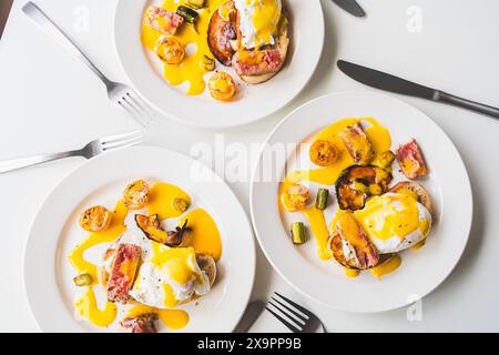 Egg Benedict with tuna, bacon, asparagus and tomatoes with hollandaise sauce on three white plates. The concept is a delicious family breakfast. Selec Stock Photo