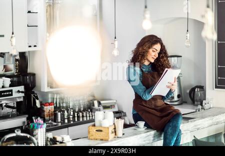 Beautiful female barista studying during part-time work in coffee shop. University student reading school notes while cafe is empty. Stock Photo