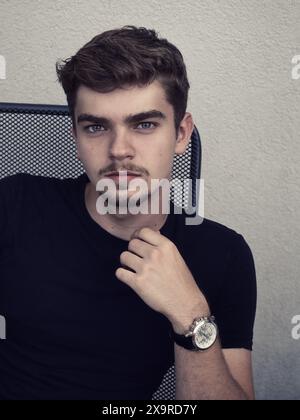 Portrait of a young man sitting outdoors against a neutral wall, wearing a black shirt and wristwatch. Casual lifestyle image with confident expressio Stock Photo