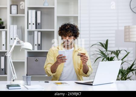 Young man worried about online banking fraud using a smartphone and credit card at a home office desk. Financial security concept. Stock Photo